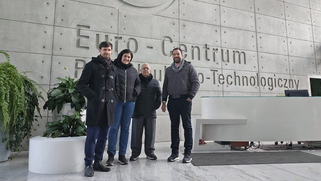 Quatro pessoas estão em pé em um hall moderno, diante de uma parede de concreto com a inscrição “Euro-Centrum Park Naukowo-Technologiczny”. Usam roupas de frio, como casacos e cachecóis. À esquerda há plantas decorativas em vasos grandes. O grupo está próximo a um balcão de recepção. Fim da descrição.