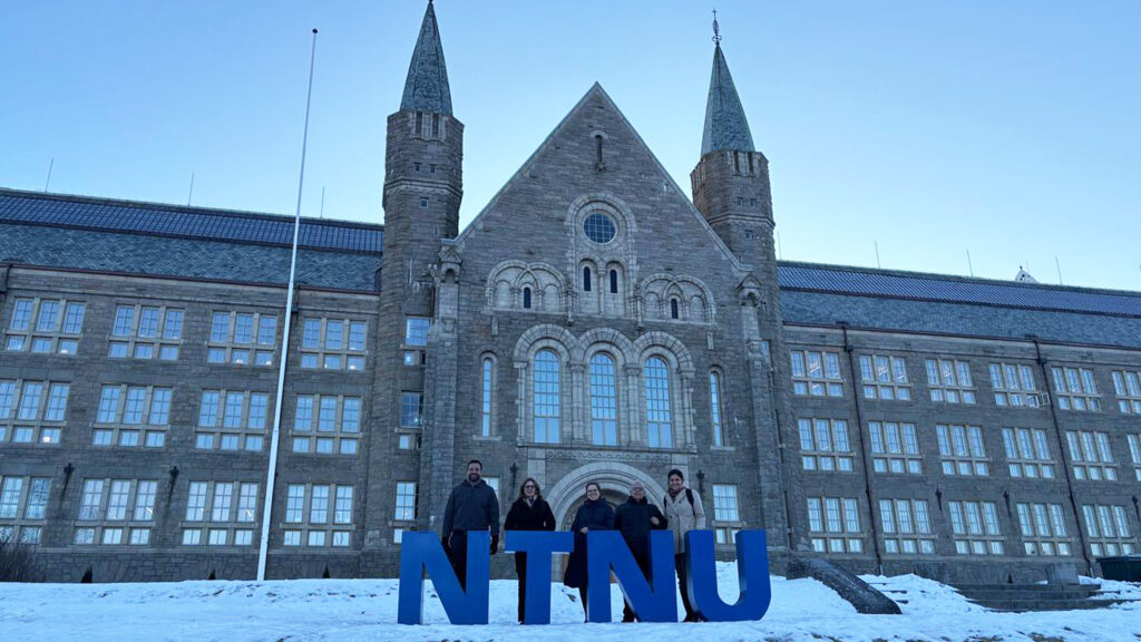 Cinco pessoas posam em frente a um grande prédio histórico de pedra, com arquitetura simétrica e duas torres laterais. No chão, há neve e letras grandes azuis formando “NTNU”. O grupo está centralizado, usando roupas de frio, e o céu está claro ao fundo. Fim da descrição.