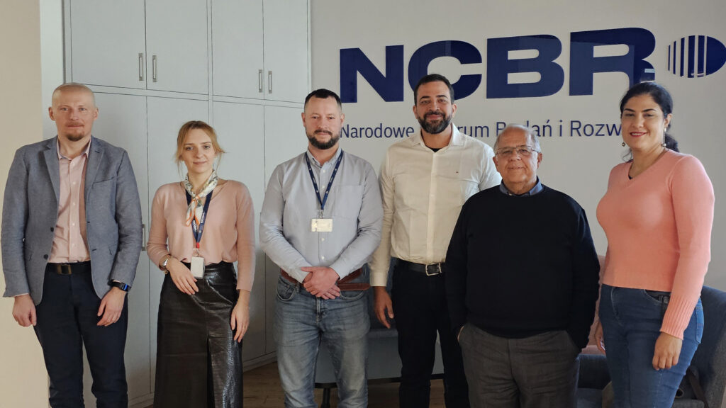 Seis pessoas posam em pé em um ambiente interno de escritório, em frente a uma parede com o logo “NCBR”. O grupo é diverso em idade e gênero, vestindo roupas formais e casuais. Ao fundo há armários brancos. Todos estão voltados para a câmera, com expressão neutra ou sorridente. Fim da descrição.