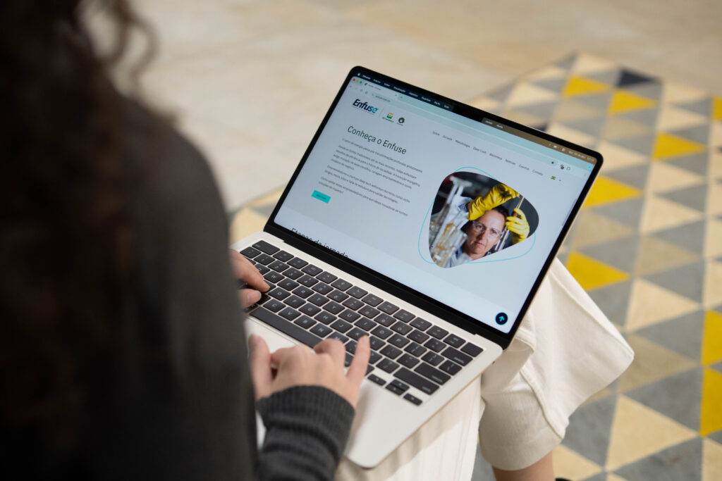 Visão por cima do ombro de uma pessoa com cabelos cacheados usando um notebook. A tela exibe o site da "Enfuse" com o título "Conheça o Enfuse" e a foto de uma cientista em um laboratório. Ao fundo, um tapete com padrão geométrico de triângulos cinzas e amarelos. Fim da descrição.