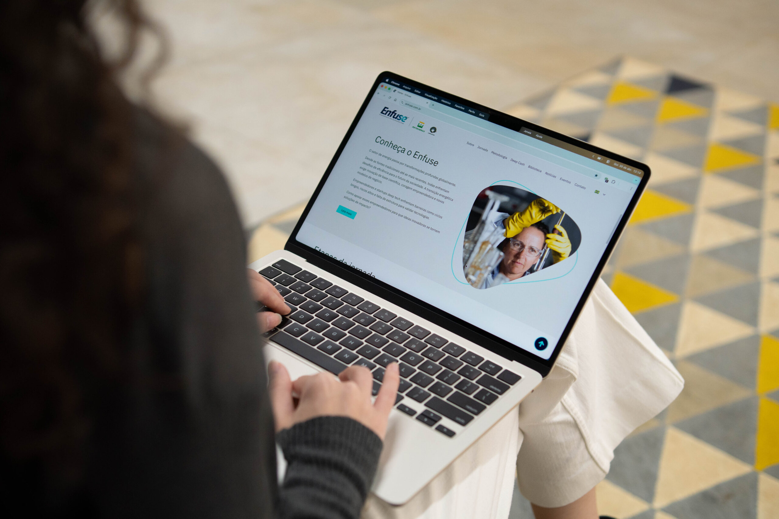 Visão por cima do ombro de uma pessoa com cabelos cacheados usando um notebook. A tela exibe o site da "Enfuse" com o título "Conheça o Enfuse" e a foto de uma cientista em um laboratório. Ao fundo, um tapete com padrão geométrico de triângulos cinzas e amarelos. Fim da descrição.