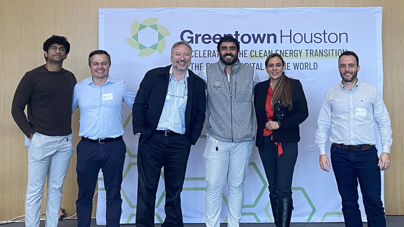 Fotografia de seis pessoas posam em frente a um painel com a marca “Greentown Houston” e a frase sobre aceleração da transição para energia limpa. O grupo está em um auditório e veste roupas sociais e casuais, registrando uma visita ao hub de inovação e energia. Fim da descrição.