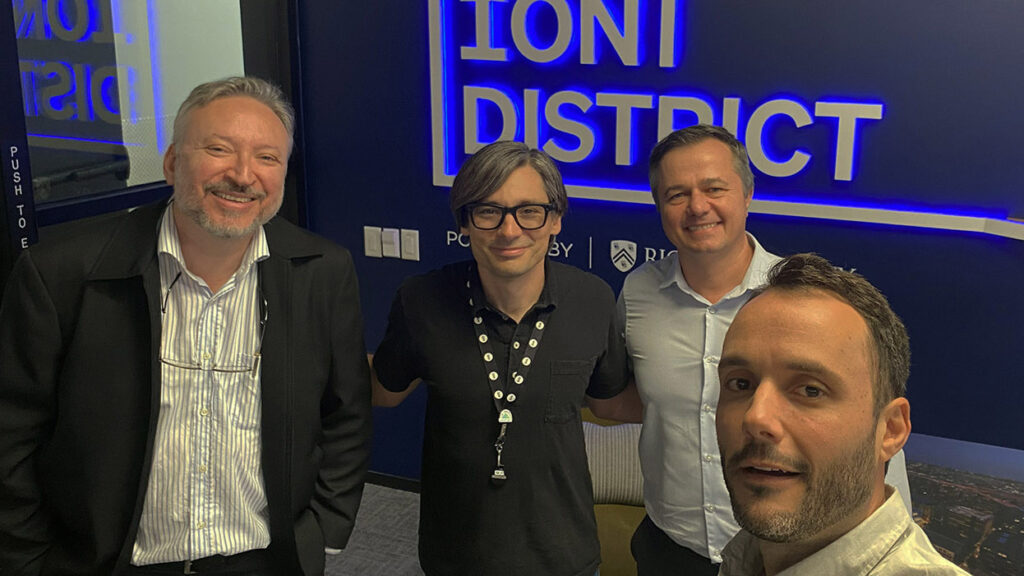 Fotografia com quatro homens tiram uma selfie em um ambiente interno diante de um letreiro iluminado com a inscrição “Ion District”. O grupo sorri para a câmera durante uma visita institucional ao ecossistema de inovação.. Fim da descrição.