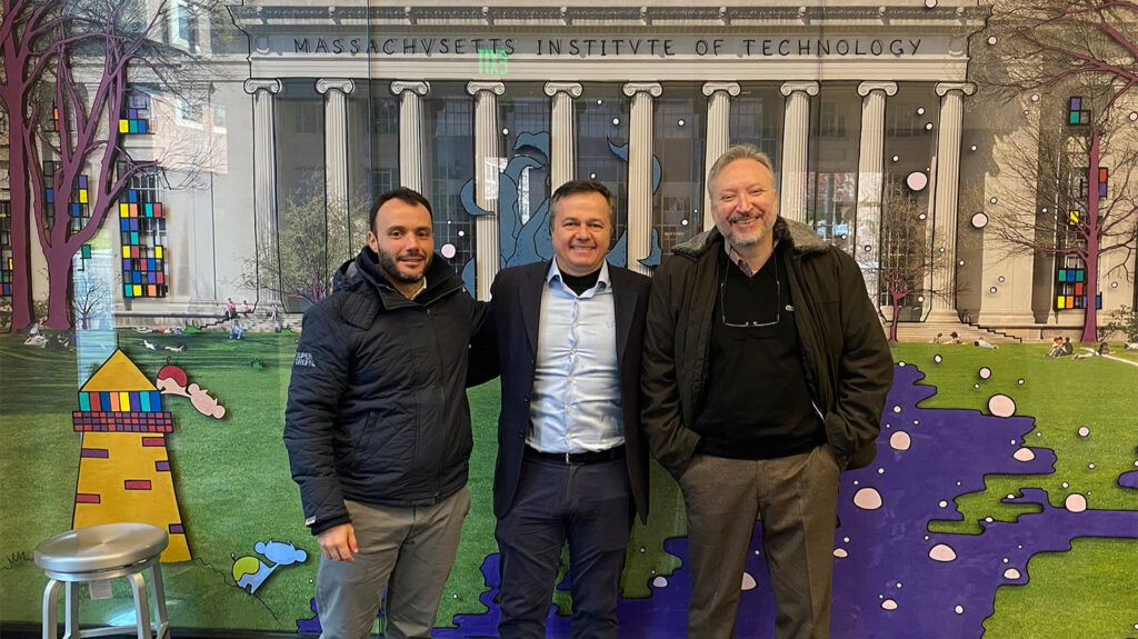 Fotografia com três homens posam sorrindo em frente a um mural ilustrado da fachada do Instituto de Tecnologia de Massachusetts (MIT). Eles estão em um ambiente interno e usam roupas sociais e casacos de inverno. Fim da descrição.
