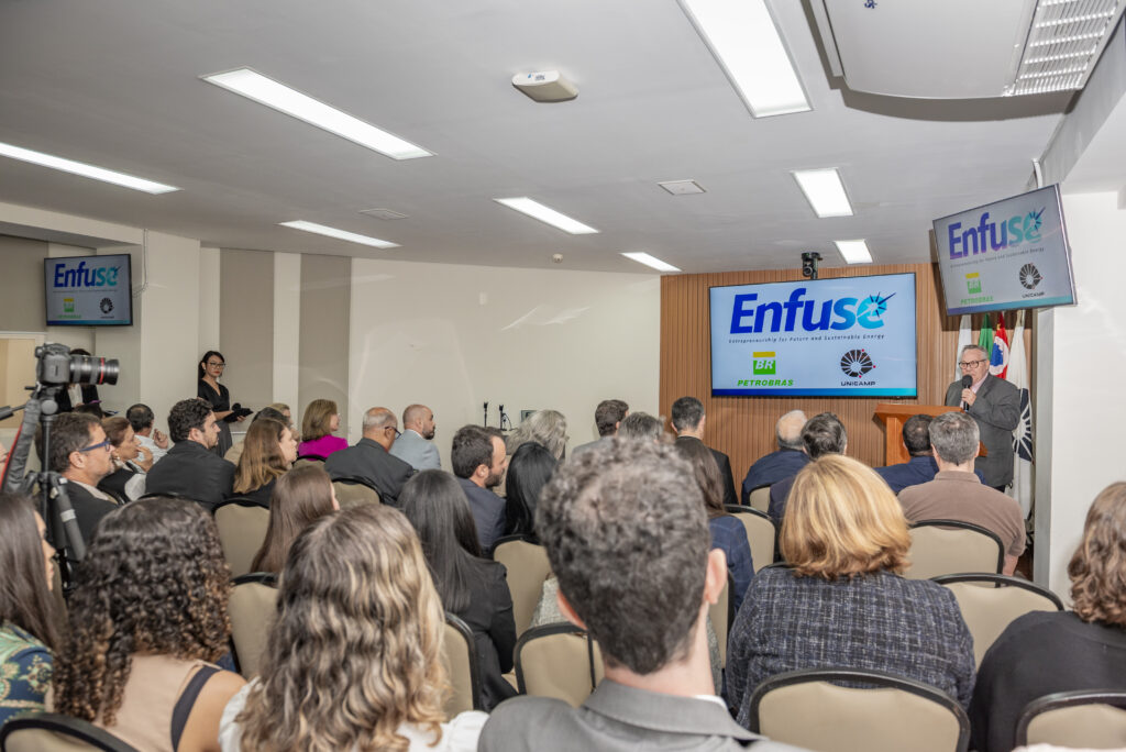 Fotografia de Sala de conferência com público sentado assistindo a um palestrante no púlpito. Ao fundo, uma tela exibe o logotipo “Enfuse” com referências à Petrobras e à Unicamp, indicando um evento institucional sobre energia sustentável. Fim da descrição.
