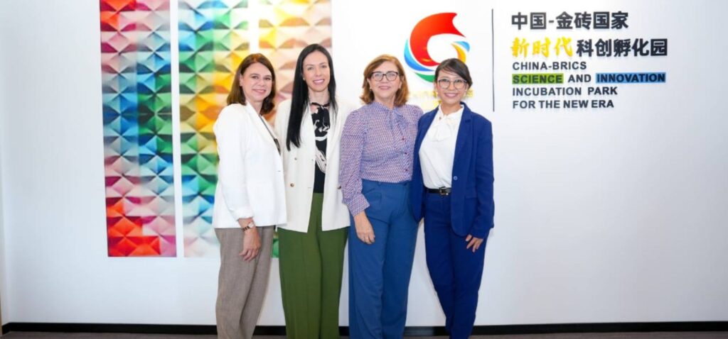 Quatro mulheres da comitiva da Inova Unicamp posam em frente a um logotipo e texto em mandarim e inglês, que dizem "CHINA-BRICS SCIENCE AND INNOVATION INCUBATION PARK FOR THE NEW ERA". A parede à esquerda tem um painel colorido abstrato. Fim da descrição.