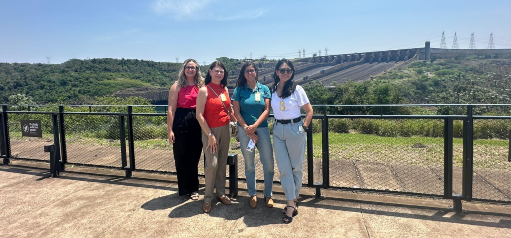As quatro mulheres da comitiva da Inova Unicamp posam em frente a Usina Hidrelétrica de Itaipu. Da esquerda para a direita, Raphaela Gomes, Mariana Zanatta Inglez, Luciana Silva e Kátia Kishi. Fim da descrição.