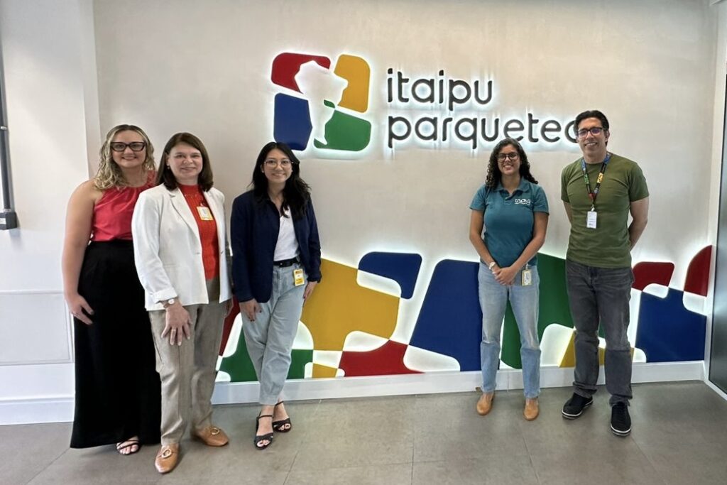 A imagem mostra a comitiva da Inova Unicamp junto com o Eduardo Bortoleto em frente a uma parede com o logo do Itaipu Parquetec. Fim da descrição.
