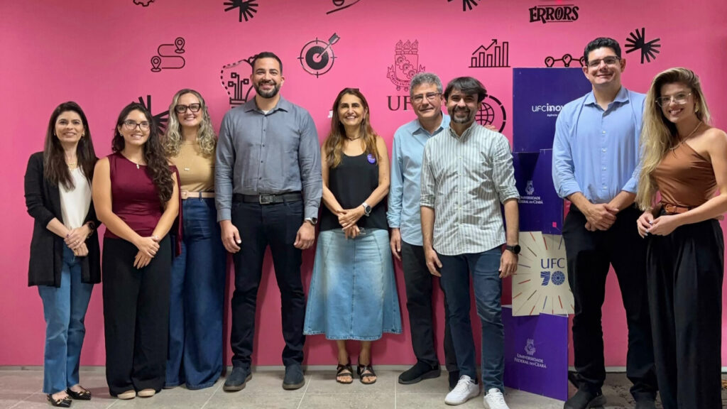 A imagem é uma fotografia de oito pessoas, pesquisadores do Enfuse e representantes da Universidade Federal do Ceará, em frente a uma parede rosa com diversos ícones e a logo da universidade. Fim da descrição.