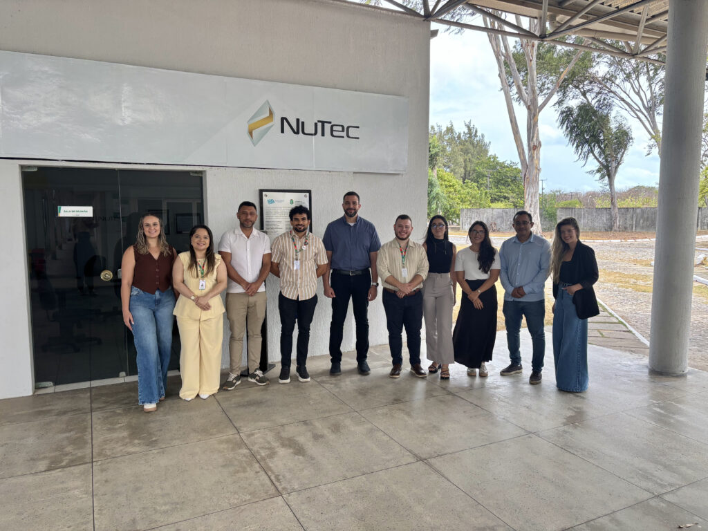A imagem é uma fotografia de dez pessoas, lado a lado, em um local coberto, próximo à uma área externa arborizada, acima delas há uma placa escrito “Nutec”. Fim da descrição.