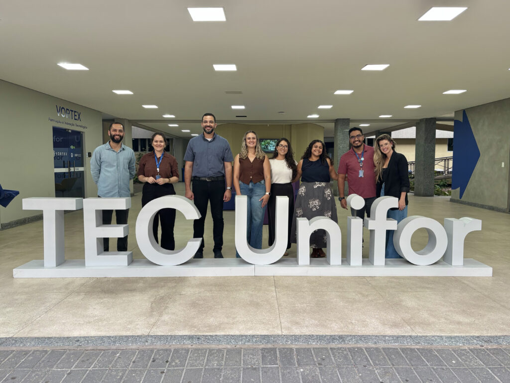 A imagem é uma fotografia de oito pessoas posando lado a lado atrás de grandes letras brancas formando a palavra ‘TEC Unifor’. A foto foi tirada em um espaço interno amplo e bem iluminado, com piso claro. Fim da descrição.