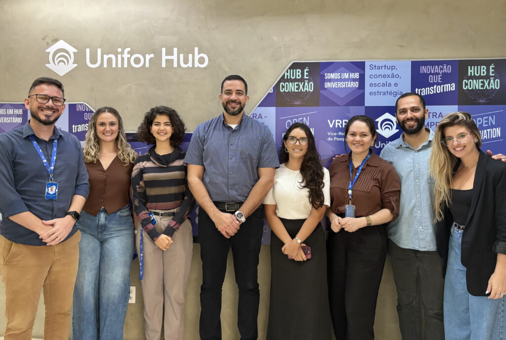 A imagem é uma fotografia de oito pessoas, lado a lado, sorrindo diante de uma parede escrito ‘Unifor Hub’, em um ambiente interno. Ao fundo há um painel com mensagens institucionais sobre inovação e conexão. Fim da descrição.