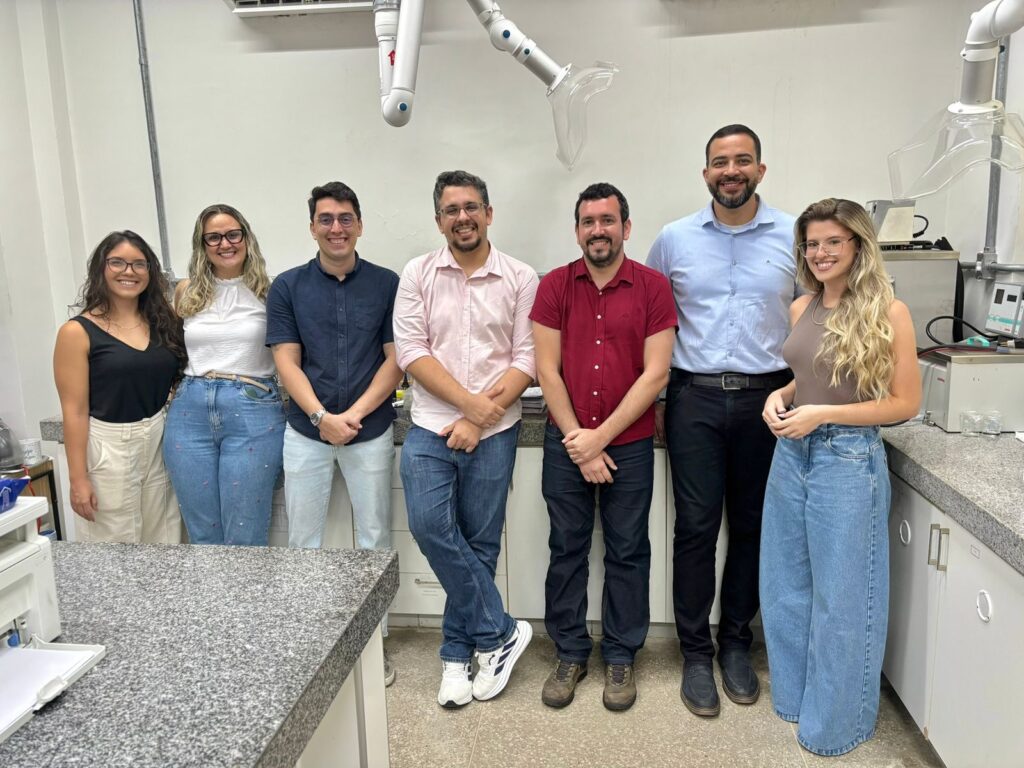 A imagem é uma fotografia de sete pessoas, lado a lado, dentro de um laboratório. O ambiente possui bancadas de granito, armários e equipamentos laboratoriais. Fim da descrição.