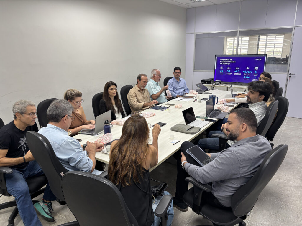 Foto de uma reunião presencial com várias pessoas sentadas ao redor de uma mesa, usando notebooks e conversando, em uma sala de reuniões. Ao fundo, há uma tela com uma apresentação sobre desenvolvimento de startups.