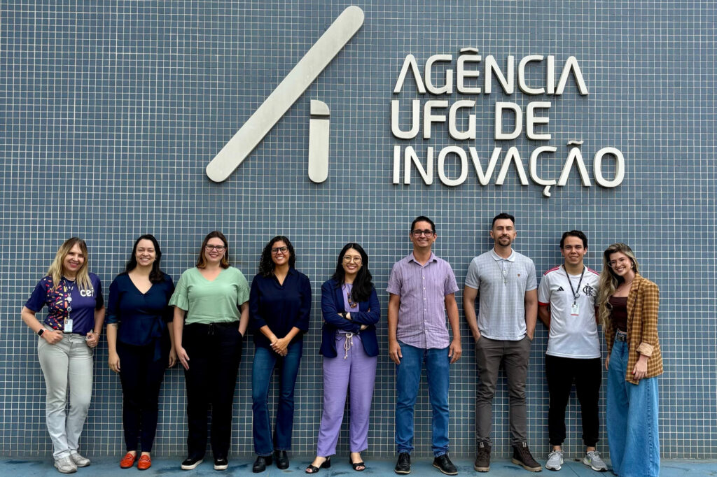 A imagem é uma fotografia de dez pessoas em pé, lado a lado, em frente a uma parede com o texto “Agência UFG de Inovação”. Fim da descrição.