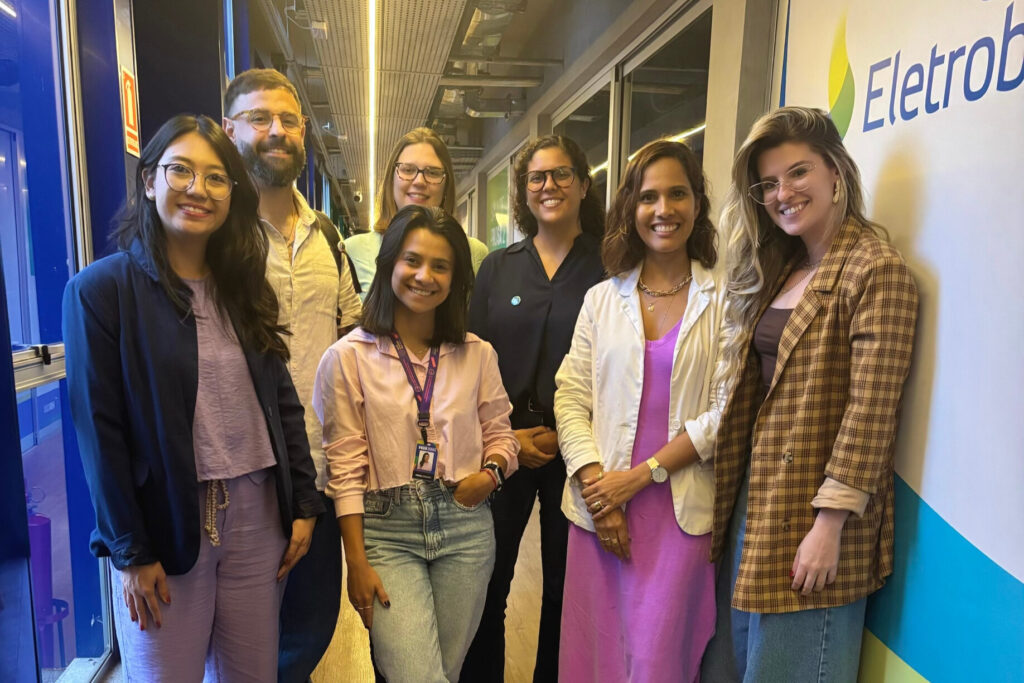 A imagem é a fotografia de oito pessoas em pé, lado a lado, em um corredor interno de um prédio, olhando para a câmera e sorrindo. Fim da descrição.