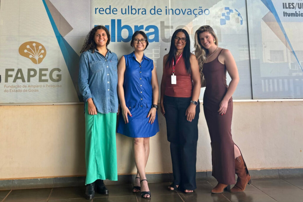 A imagem é uma fotografia de quatro mulheres em pé, lado a lado, em frente a um painel com o texto “rede ulbra de inovação”. Fim da descrição.