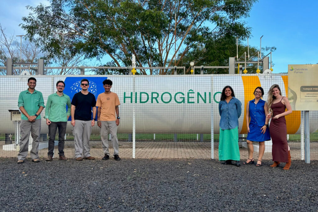 A imagem é uma fotografia de oito pessoas em pé, lado a lado, em área externa, em frente a uma estrutura com a palavra “Planta de Hidrogênio”. Fim da descrição.