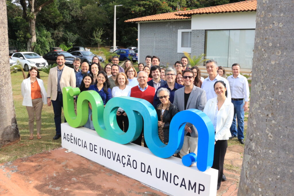 Grupo de pessoas posando atrás do letreiro ‘Inova – Agência de Inovação da Unicamp’, em área externa do campus. Fim da descrição.