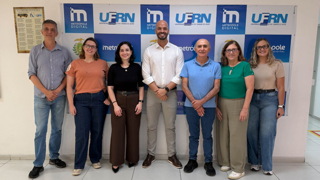 Fotografia de um grupo de sete pessoas em pé, lado a lado. Eles estão em um ambiente interno, em frente a um painel que exibe repetidamente os logotipos do "Metrópole Digital" e da "UFRN". Fim da descrição.