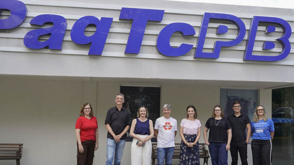 Grupo de oito pessoas, lado a lado em frente à fachada de um prédio com a grande placa "PaqTcPB" em letras azuis na parte superior. Fim da descrição.