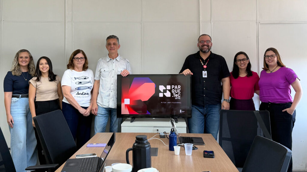 Grupo de sete pessoas posando lado a lado em uma sala de reunião. Ao centro, há uma televisão sobre um móvel exibindo a tela com o texto "Parque.TeC UFPE". O ambiente é claro, com paredes brancas e iluminação uniforme. Fim da Descrição.