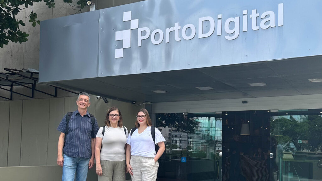 Três pessoas em frente à entrada de um prédio com a placa "Porto Digital" na parte superior. Ao fundo, há portas de vidro que permitem ver o interior do edifício. Fim da descrição.