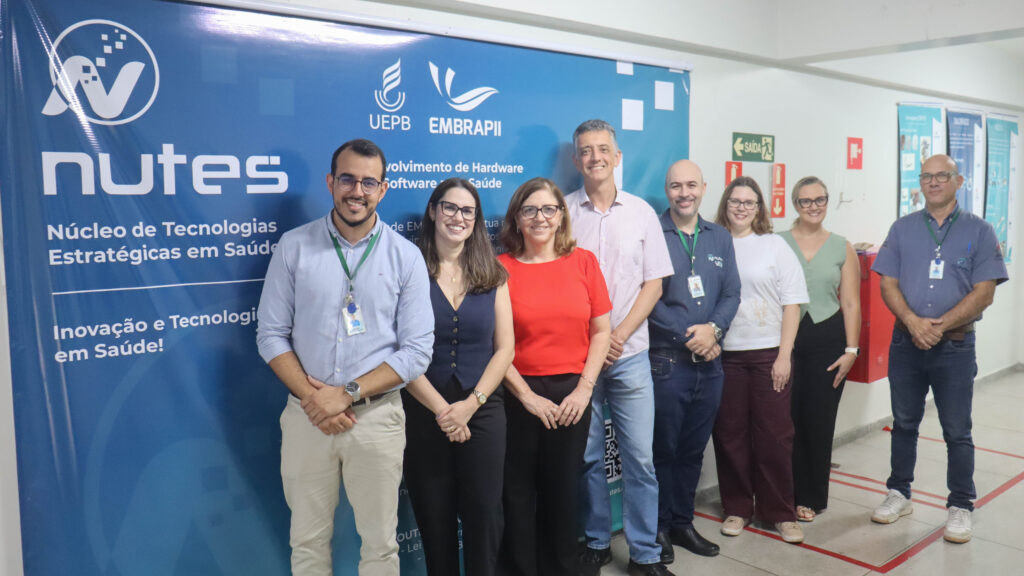 Grupo de oito pessoas, lado a lado em um corredor interno. Ao fundo, há um grande painel azul com os logotipos "NUTES", "UEPB" e "EMBRAPII". Fim da descrição.