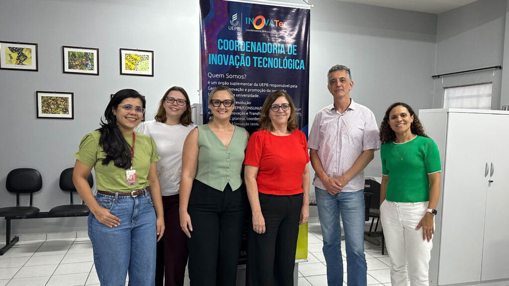 Grupo de seis pessoas, lado a lado em uma sala. Ao fundo, há um banner com os logotipos "UEPB" e "InovaTec". O ambiente tem paredes claras, quadros decorativos, cadeiras encostadas na parede e um armário branco, com iluminação interna uniforme. Fim da descrição.