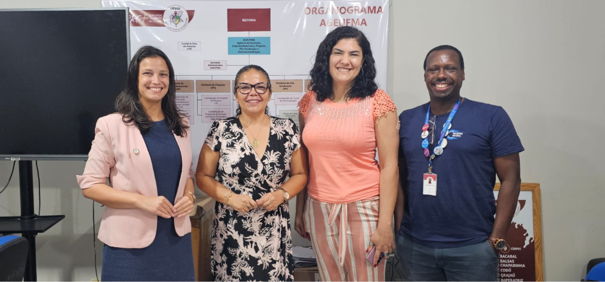 Foto de quatro pessoas em pé, sorrindo para a câmera, em um ambiente institucional. Ao fundo, há um painel com um organograma e logotipos, indicando um contexto acadêmico ou administrativo. Fim da descrição.